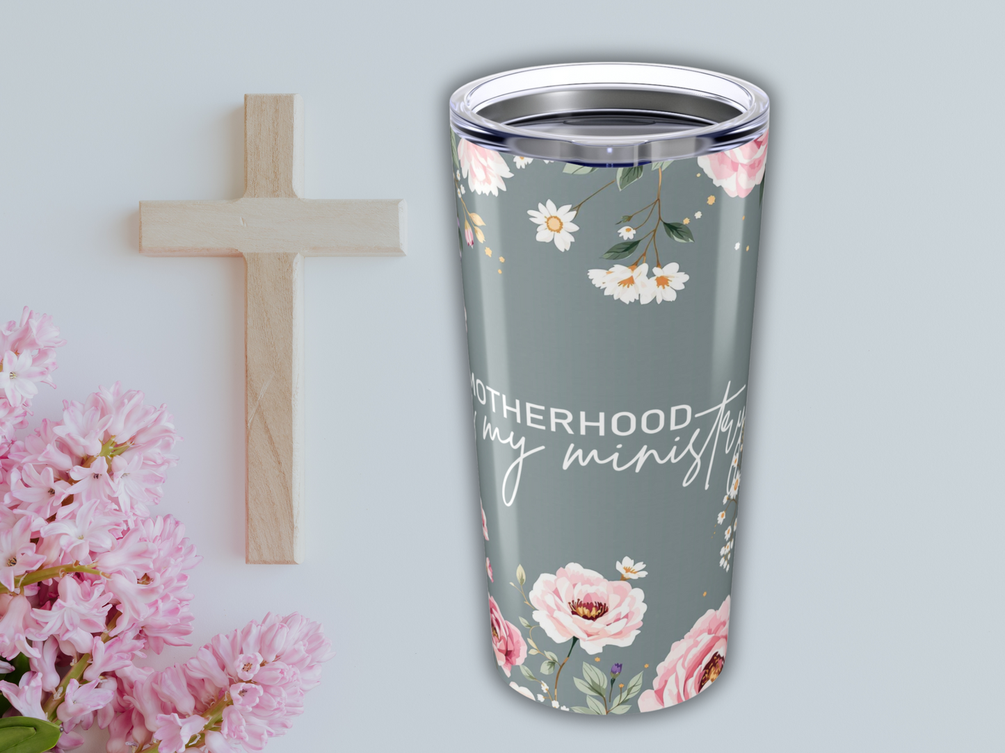 A floral travel mug with the text "Motherhood is my ministry" is placed beside a wooden cross and pink flowers on a light gray surface.