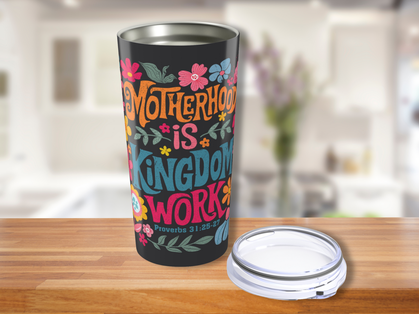 Colorful travel mug with floral design and text, "Motherhood is Kingdom Work - Proverbs 31:25-27," on a wooden counter in a bright kitchen.