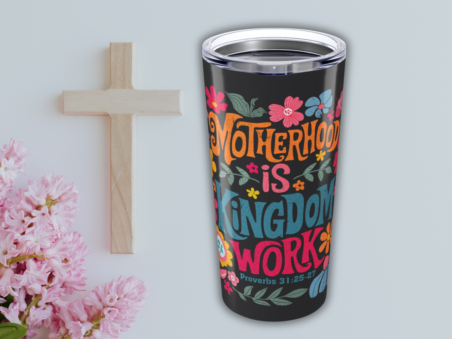 A wooden cross and pink flowers are placed beside a black 20oz Stainless Steel Tumbler. The tumbler features colorful floral designs and the text "Motherhood is Kingdom Work" with a Bible verse.