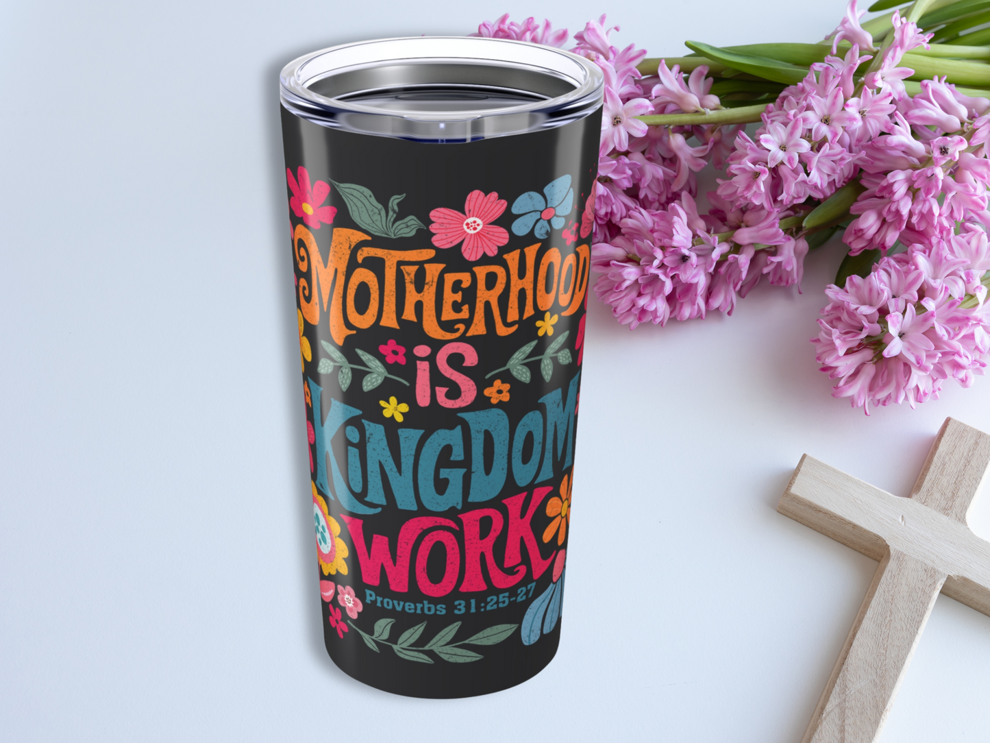 A colorful 20oz Stainless Steel Tumbler with "Motherhood is Kingdom Work" in vibrant lettering, surrounded by flowers. Nearby are pink hyacinths and a wooden cross, conveying a serene, spiritual tone.