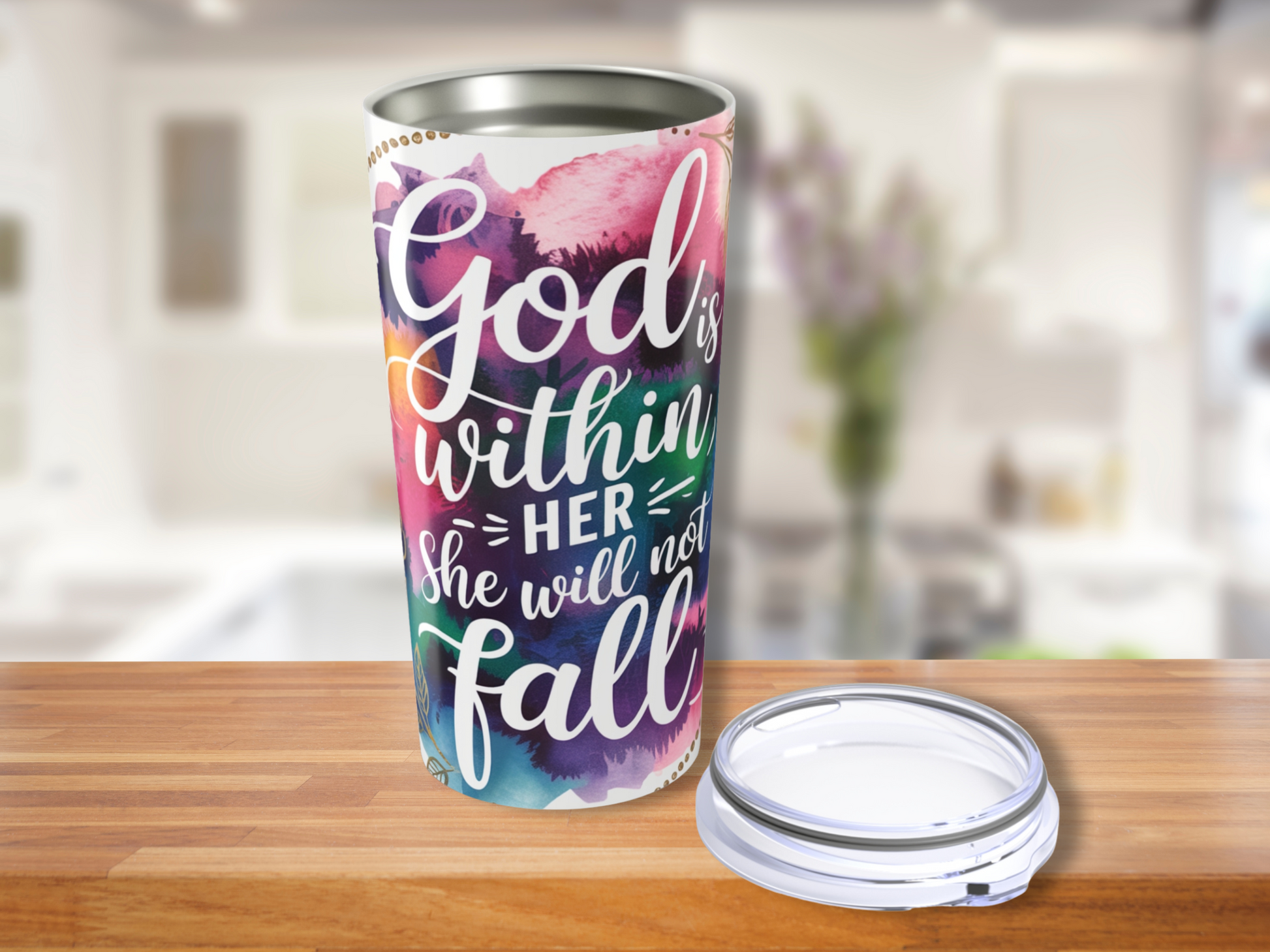 A colorful travel mug with the inspirational text "God is within her, she will not fall" on a blurred kitchen background. A lid is placed beside it.