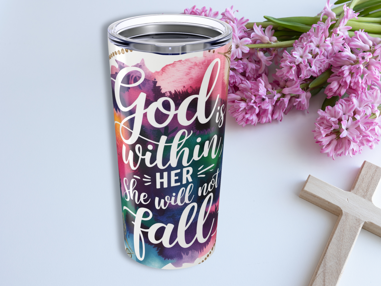A colorful religious tumbler with the text "God is within her, she will not fall" is beside pink flowers and a wooden cross, conveying faith and positivity.