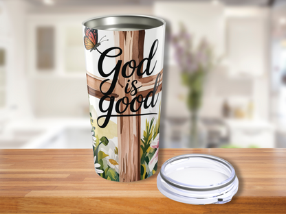 Stainless Steel Tumbler with "God is good" text over a wooden cross, adorned with a butterfly and flowers. Background shows a blurred, bright kitchen.