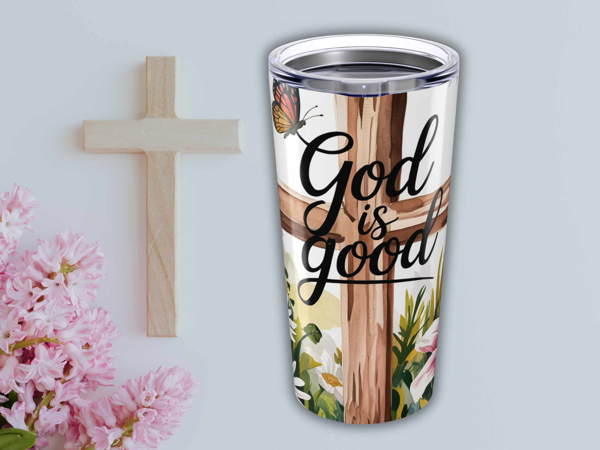 Tumbler with "God is good" text, floral design, and butterfly, next to a wooden cross and pink flowers. Calm, uplifting spiritual theme.