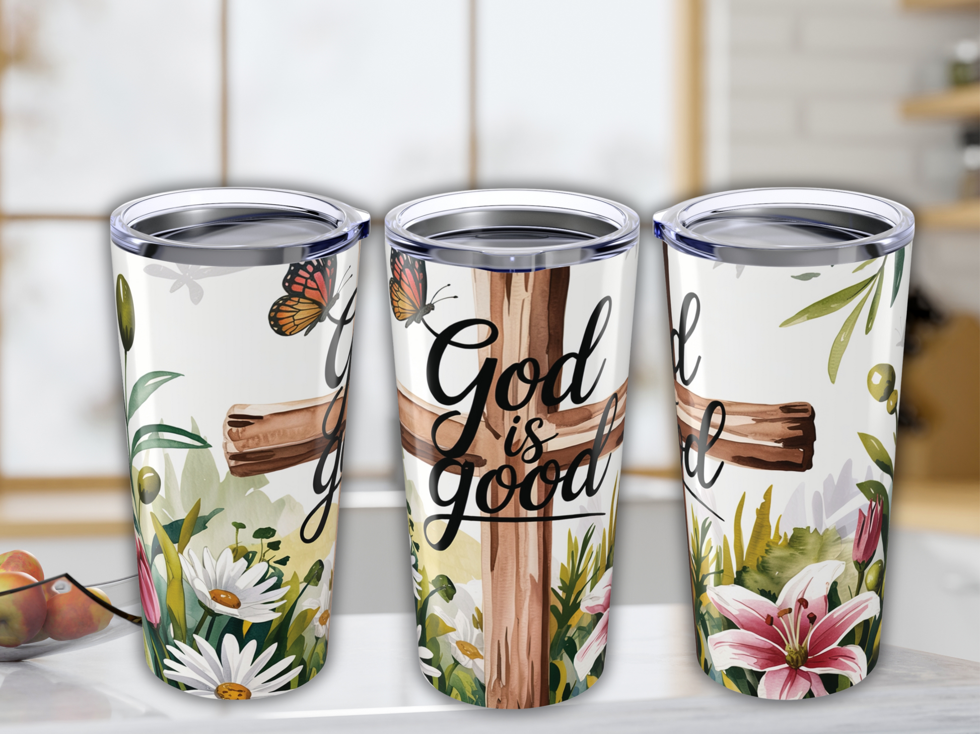 Three tumbler cups with floral designs featuring daisies and lilies. A wooden cross with the words "God is good" and butterflies adorn the center cup.
