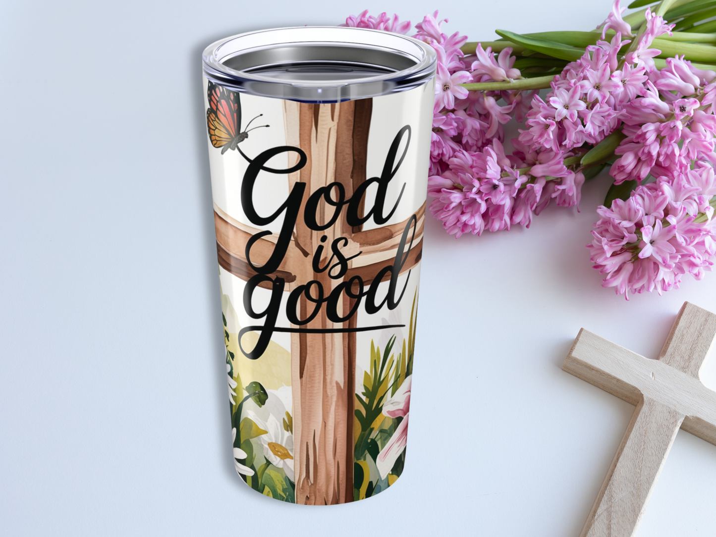 Tumbler with a floral cross design and "God is good" text. A butterfly is perched on top. Pink flowers and a wooden cross lie beside. Inspirational tone.