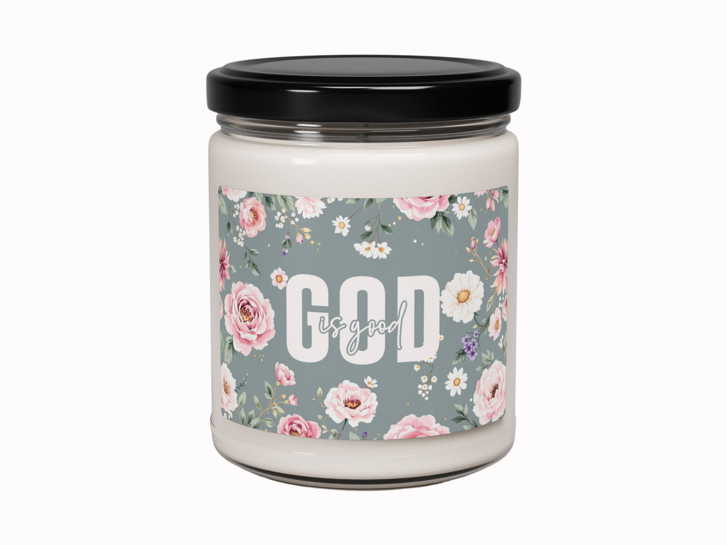 A white candle in a glass jar with a black lid. The label features pink and white flowers on a blue background, with the text "God is good" in white.