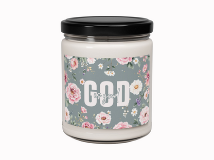 A white candle in a glass jar with a black lid. The label features pink and white flowers on a blue background, with the text "God is good" in white.
