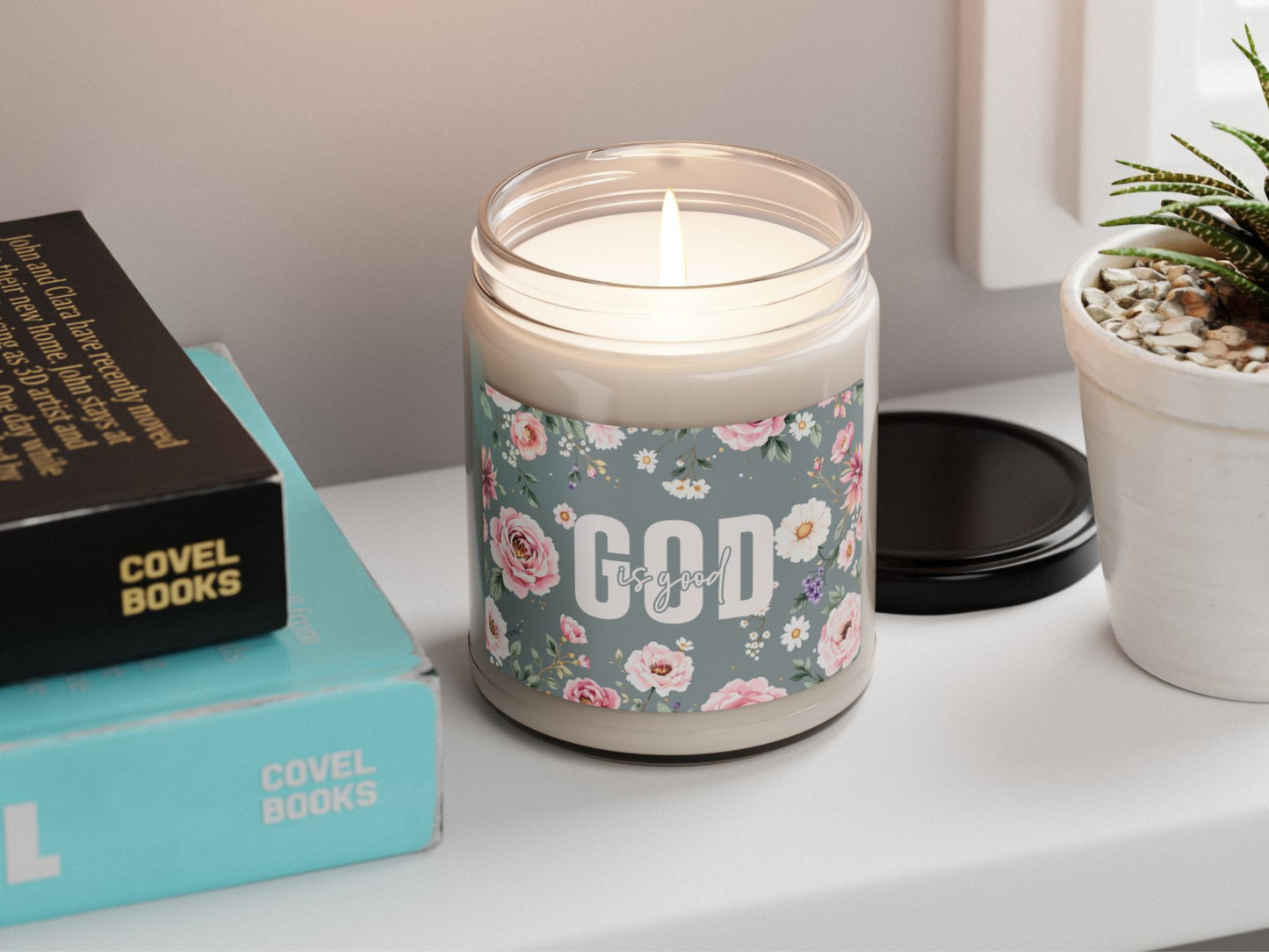 A lit candle with floral "God is good" label sits on a shelf. Nearby are stacked books with "Covel Books" visible. A potted plant adds greenery. Warm, cozy ambiance.