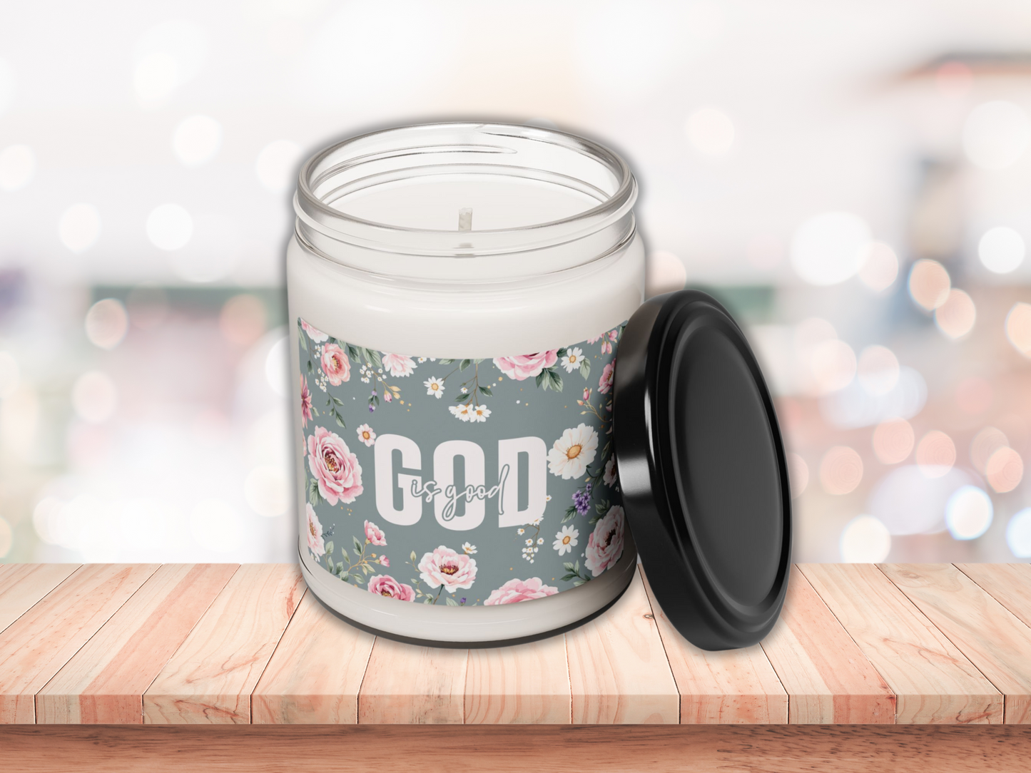 A floral-patterned candle jar on a wooden surface with "God is good" written in elegant script. The black lid is placed beside the jar, with soft bokeh lights in the blurred background, creating a warm, inviting atmosphere.