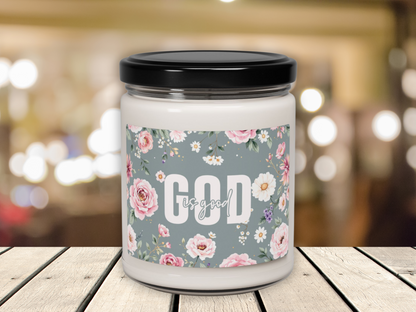 A white candle with a black lid sits on a wooden surface. Its label features pink and white flowers on a gray background with "God is good" text. The background shows softly blurred lights, adding a cozy, serene atmosphere.