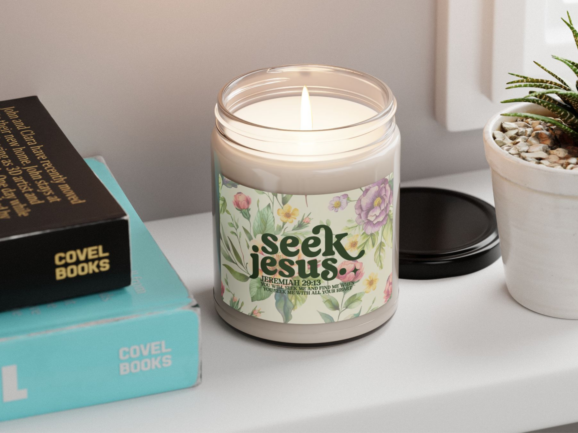 Lit candle in a glass jar with floral label reading "Seek Jesus, Jeremiah 29:13." Nearby are books and a white plant pot with stones. Peaceful atmosphere.