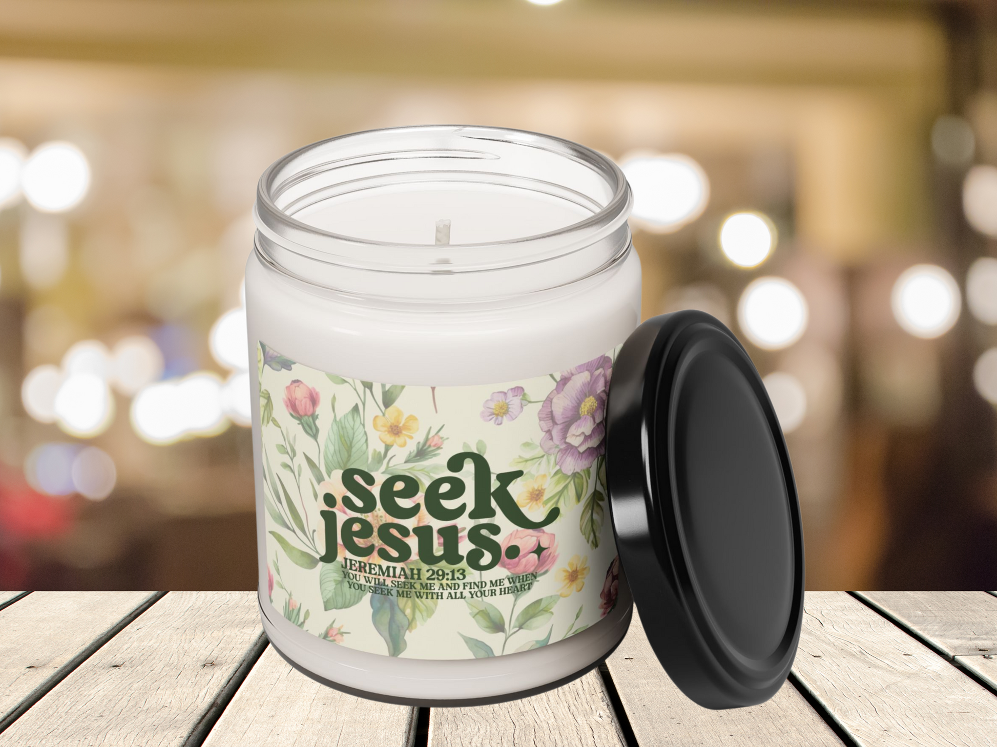 A white candle jar with a floral label reading "Seek Jesus," featuring Jeremiah 29:13. The background is softly lit with bokeh lights, creating a warm ambiance.