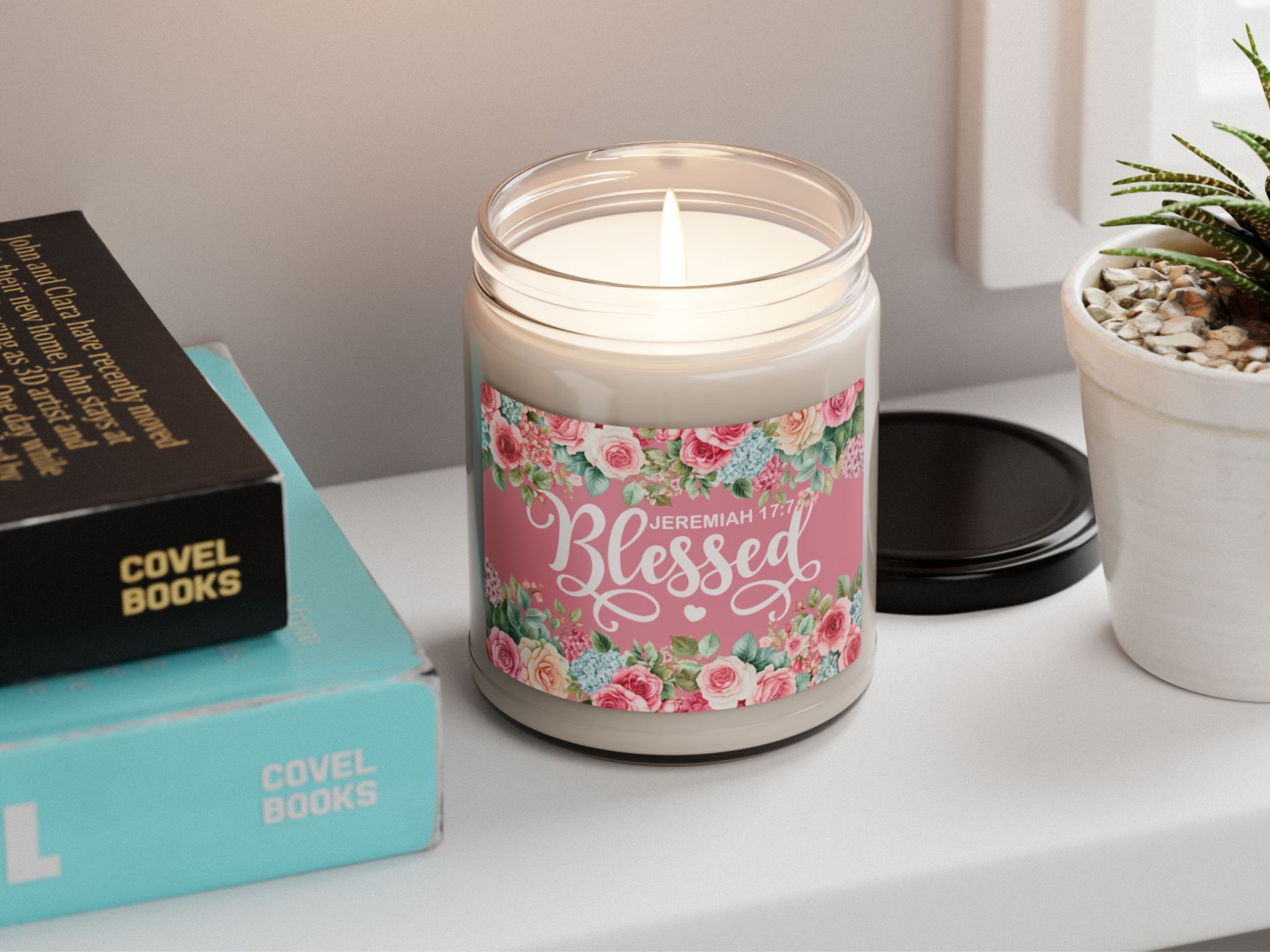 A lit candle jar with floral design and "Blessed Jeremiah 17:7" text sits on a shelf beside books and a potted plant, exuding a cozy, peaceful vibe.