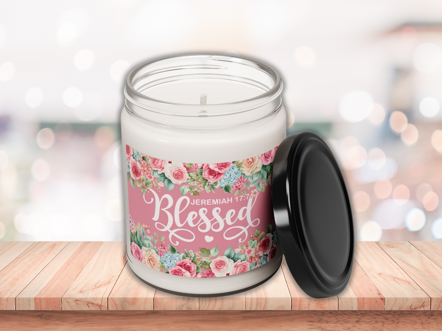 A white candle in a glass jar sits on a wooden surface. The label reads "Blessed" with floral designs. The jar's lid is black and placed beside it.