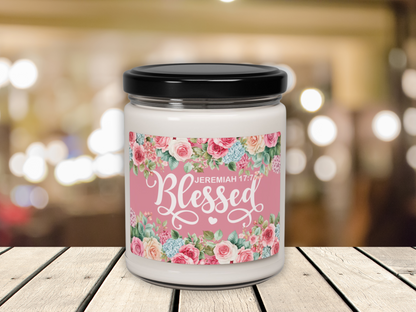 A white candle in a jar with a black lid, featuring a floral label with roses and "Blessed Jeremiah 17:7" text, set on a wooden table with blurred warm lights in the background.