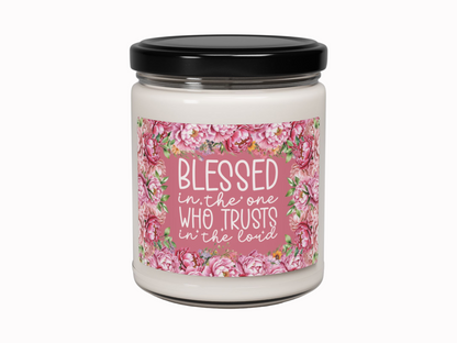 A white candle in a glass jar with a black lid. The label reads, "Blessed in the one who trusts in the Lord," surrounded by pink floral designs.