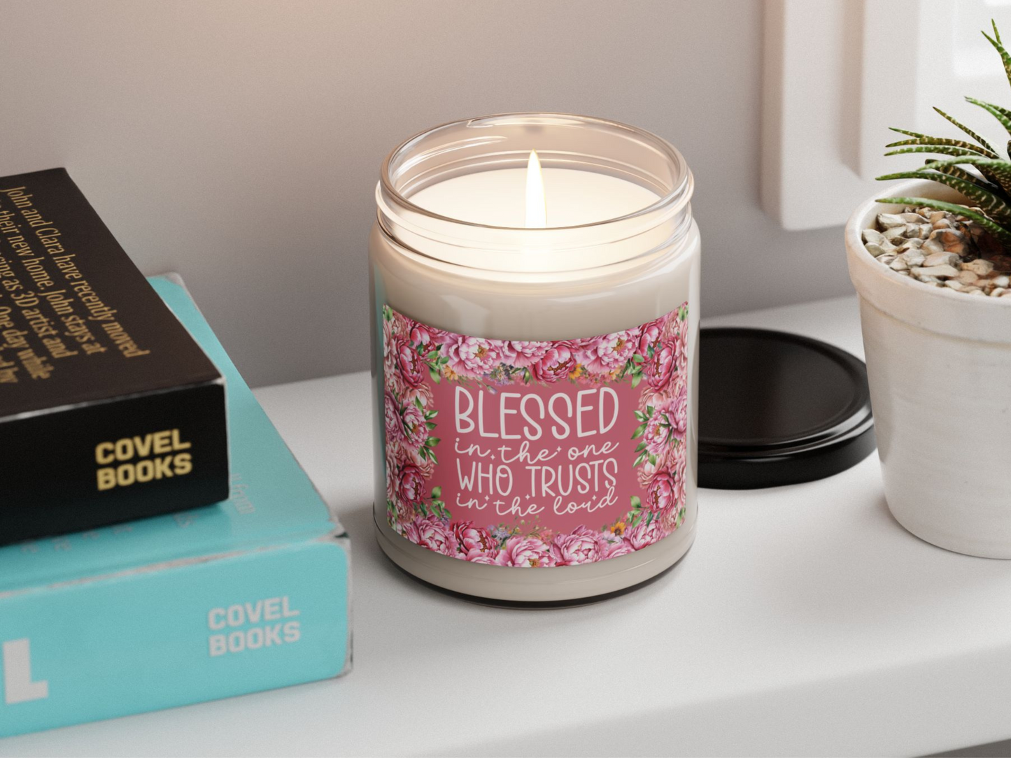 A lit candle in a jar with a floral label reading "Blessed is the one who trusts in the Lord" rests on a table beside stacked books and a small plant.