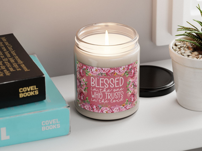 A lit candle in a jar with a floral label reading "Blessed is the one who trusts in the Lord" rests on a table beside stacked books and a small plant.