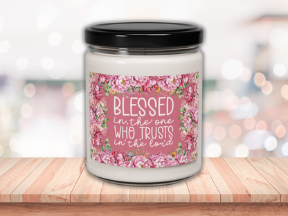A white candle jar with a black lid sits on a wooden surface. The label features pink peonies framing the words, "Blessed is the one who trusts in the Lord." The background is softly blurred with warm bokeh lights, giving a serene and uplifting feel.