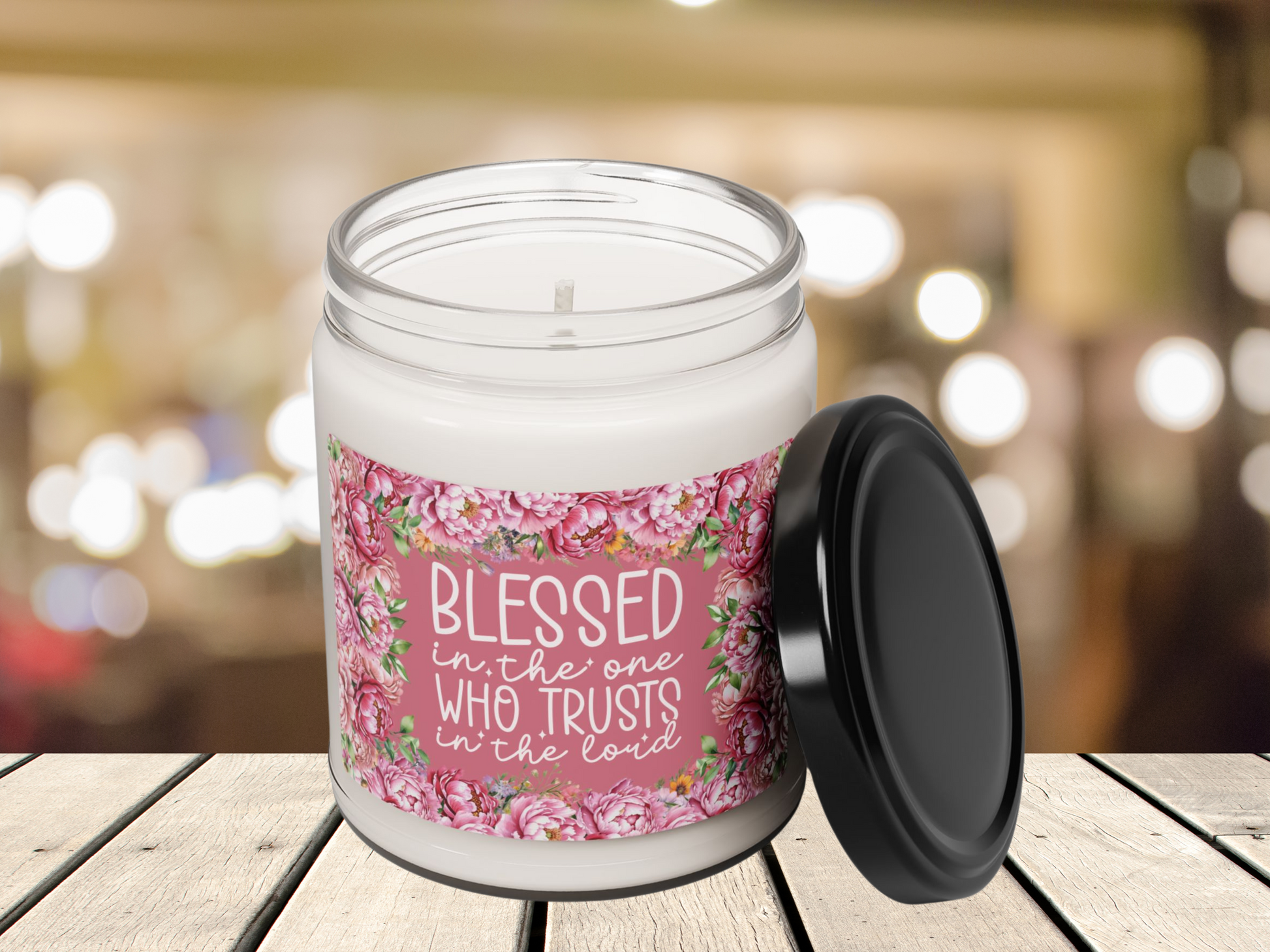 A white candle in a glass jar on a wooden surface. The floral label reads "Blessed in the one who trusts in the Lord." Background is softly blurred lights.