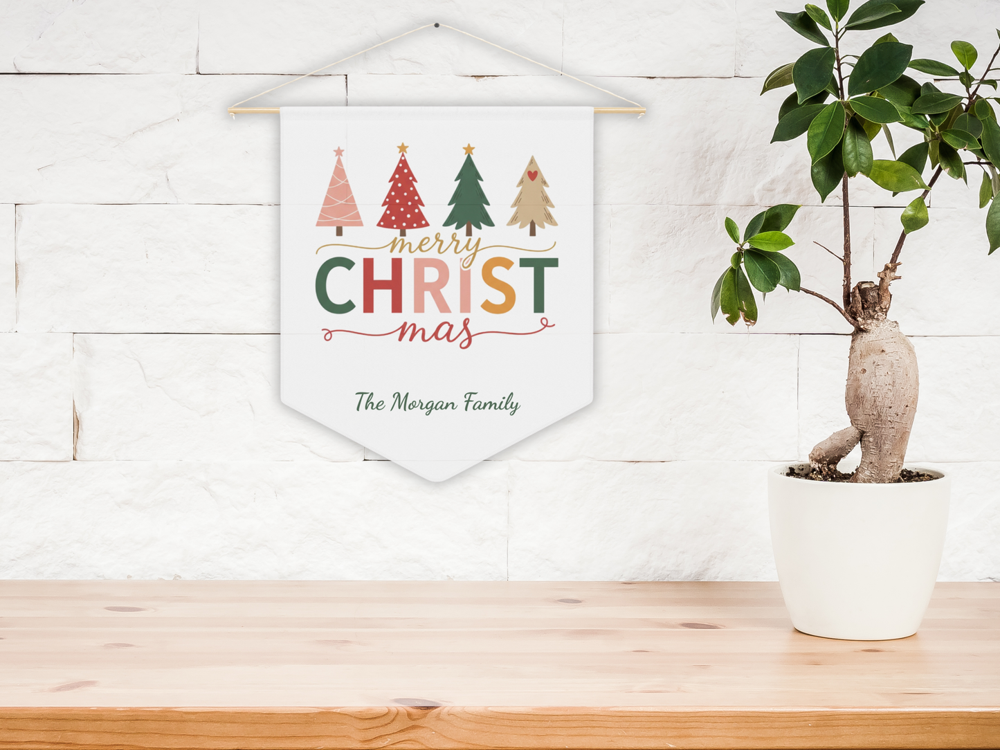 Decorative Christmas banner with trees and 'Merry Christmas' text on a white wall with a plant in the foreground.