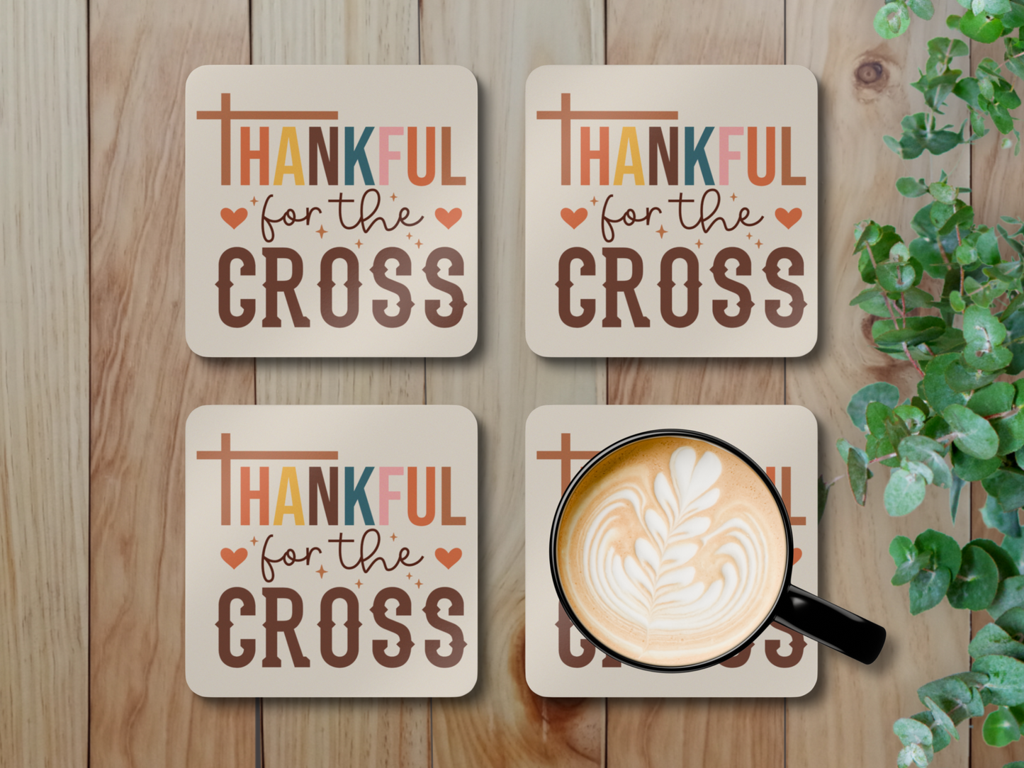 Set of four “Thankful for the Cross” coasters with latte mug styled on rustic wooden surface with greenery.