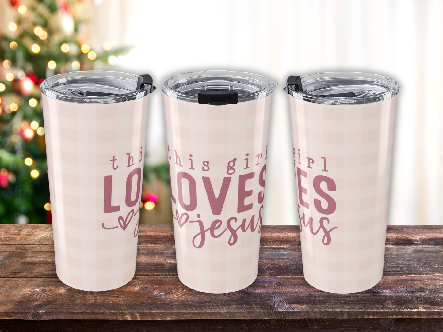 Three tumblers with 'this girl loves Jesus' text on a wooden surface with a blurred Christmas tree background.