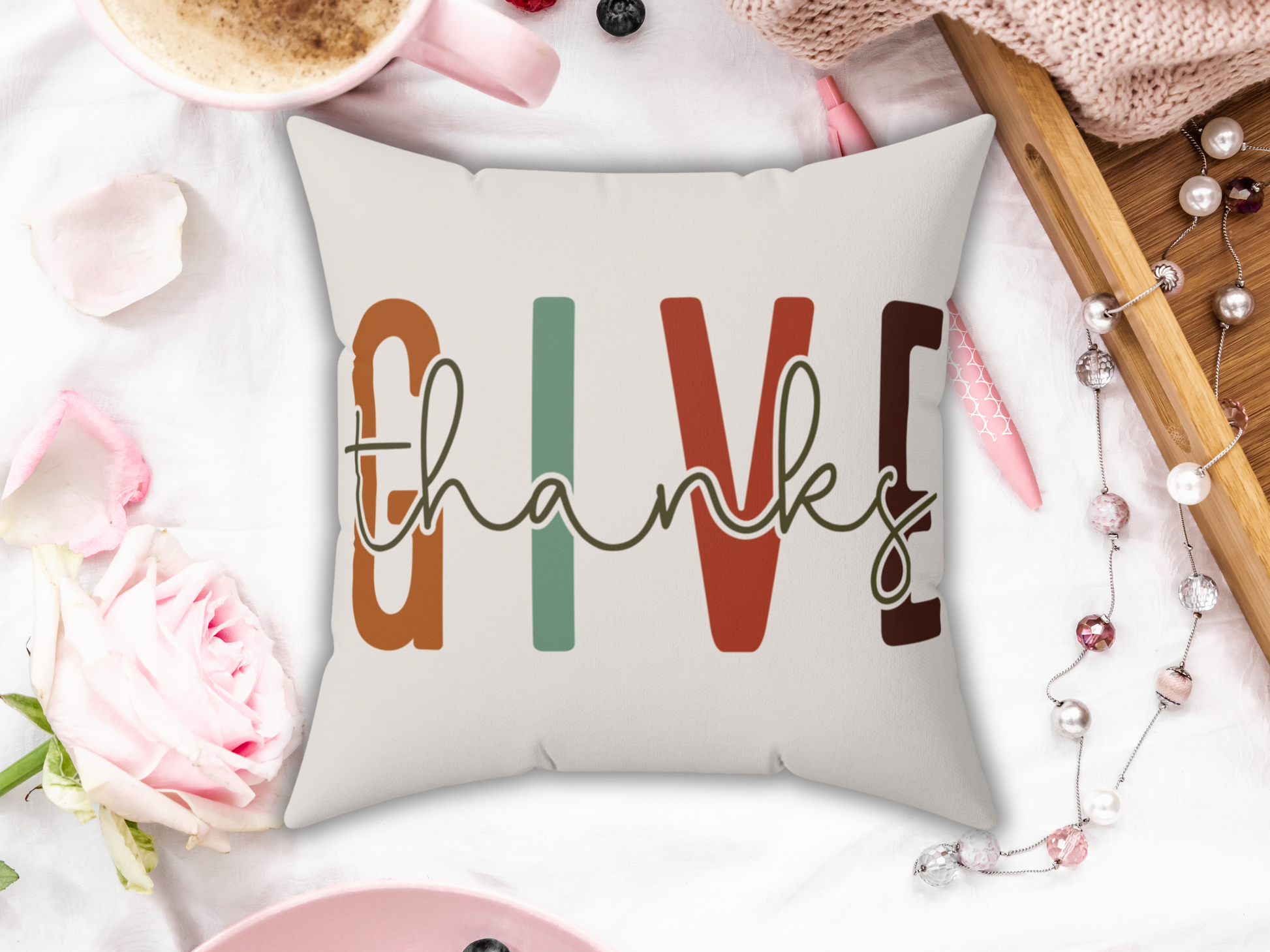 A decorative pillow with the word "thanks" in cursive, overlaying "GIVE" in bold autumn colors. Surrounded by a rose, coffee, and cozy textiles.