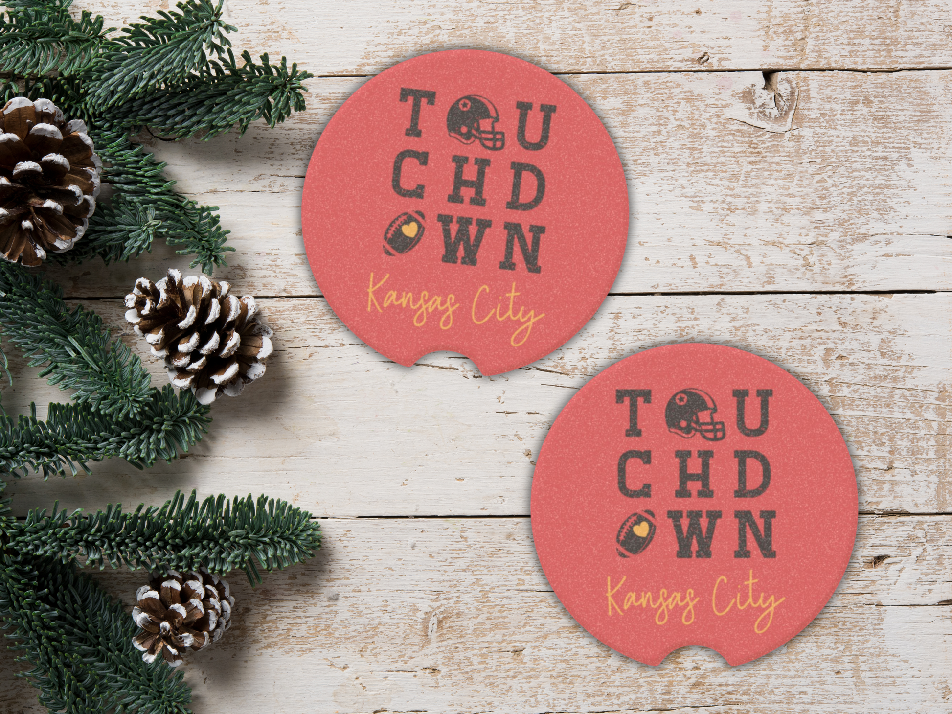 Two red 'Touchdown Kansas City' coasters on a wooden surface with pine cones and greenery.