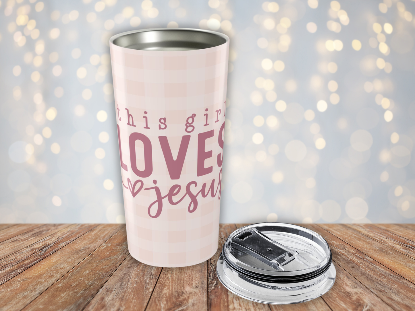 Pink plaid tumbler with 'this girl loves Jesus' text on a wooden surface with a blurred light background