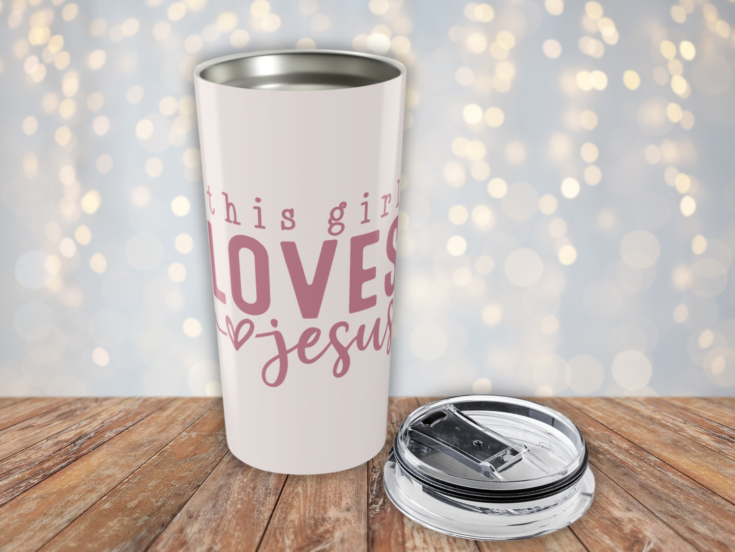 Pink tumbler with pink text 'This girl loves Jesus' on a wooden surface with a blurred light background