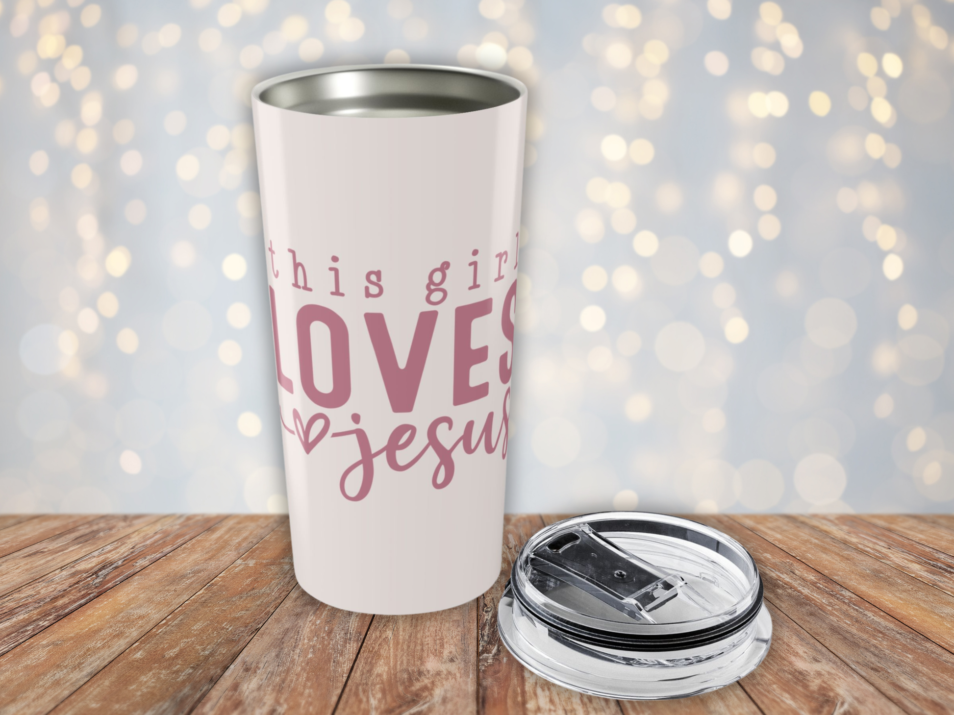 Pink tumbler with pink text 'This girl loves Jesus' on a wooden surface with a blurred light background