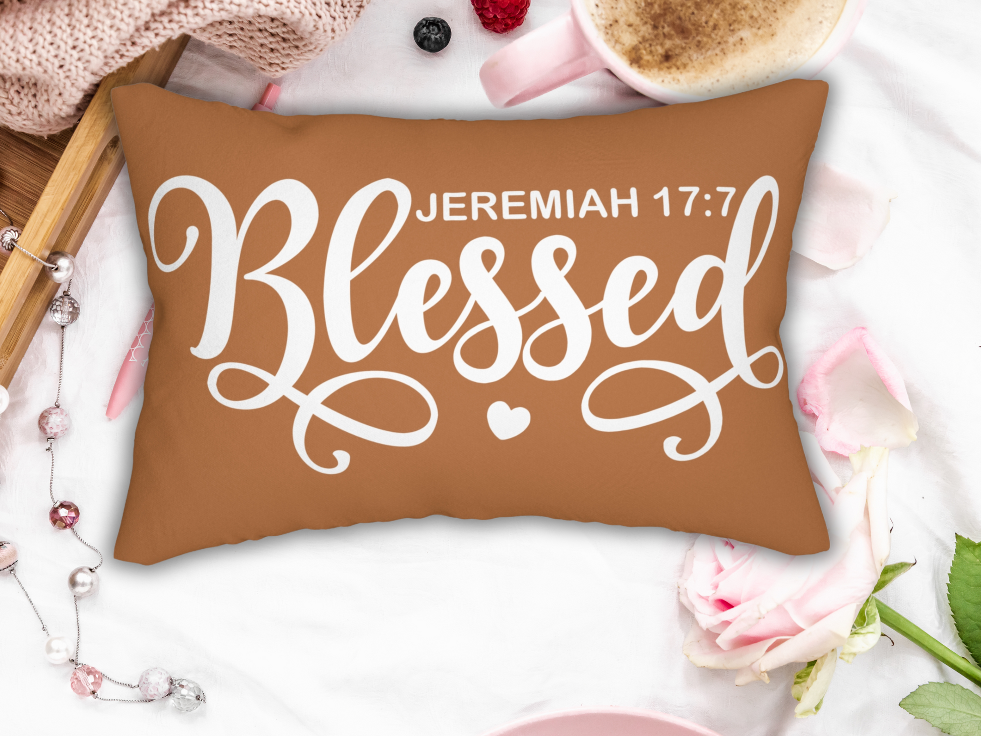 Scripture pillow on a white surface with pink mug, rose, knit blanket, and pearls for a soft cozy vibe.