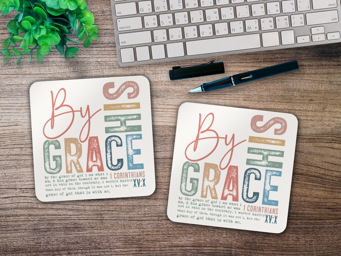 Pair of scripture coasters with “By His Grace” design styled on a wooden desk with keyboard, pen, and greenery.