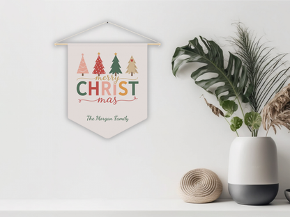 Decorative Christmas pennant with colorful text and trees, hanging on a white wall with plants and a vase below.