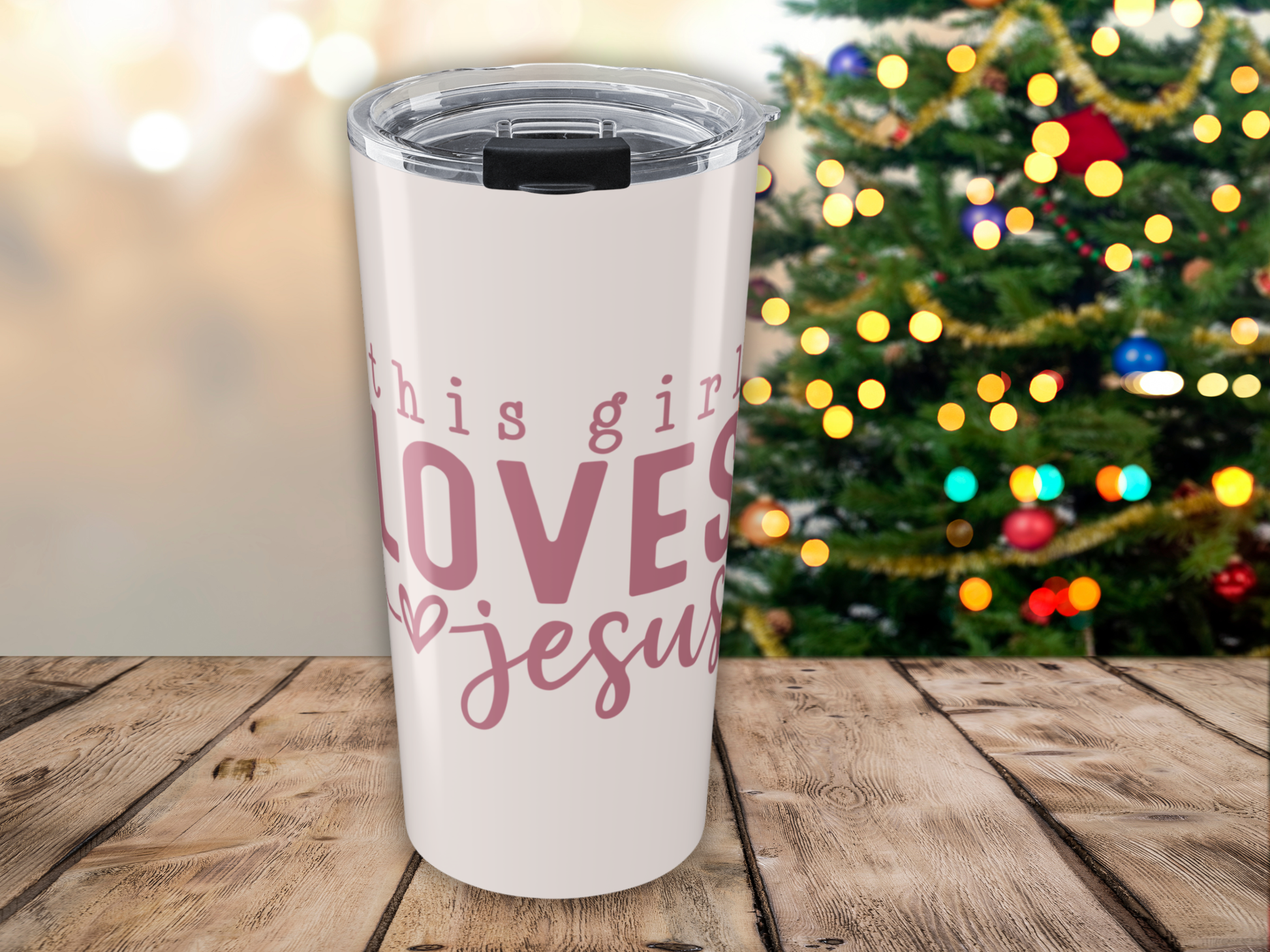 Pink tumbler with 'this girl loves Jesus' text in pink, placed on a wooden surface with a Christmas tree in the background.