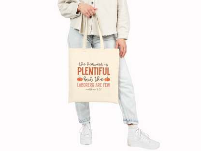 Person holding a beige tote bag with the text, "the harvest is PLENTIFUL but the laborers are few, Matthew 9:37," accented by small pumpkin drawings. Casual attire.