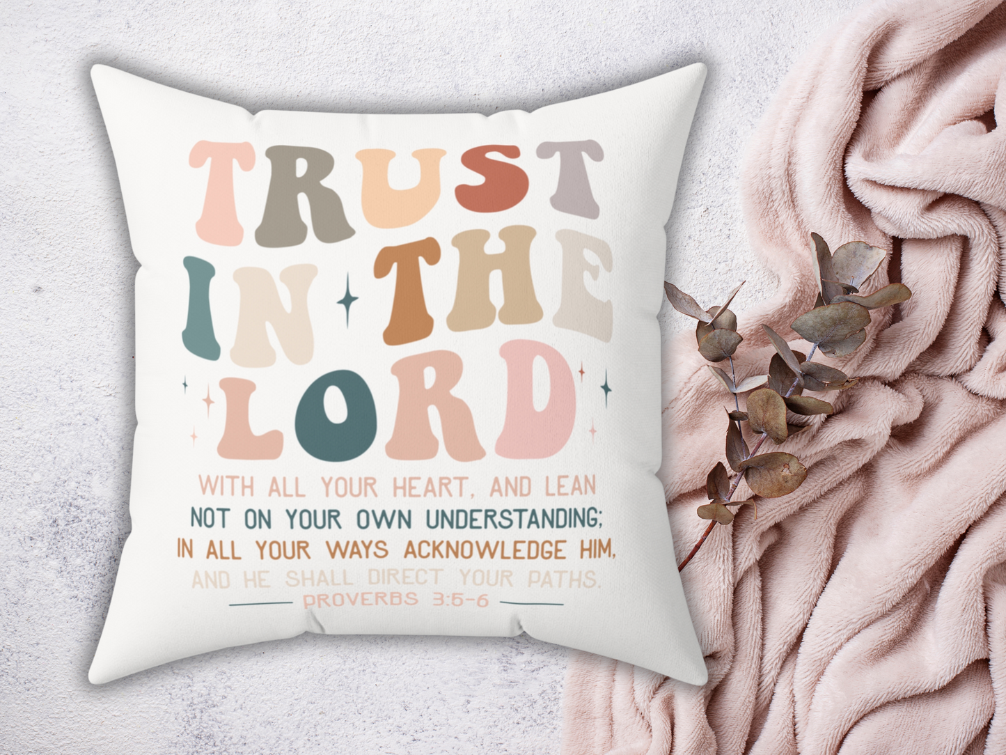 White scripture pillow with Proverbs 3:5-6 “Trust in the Lord” text, styled with blush pink blanket and eucalyptus leaves.