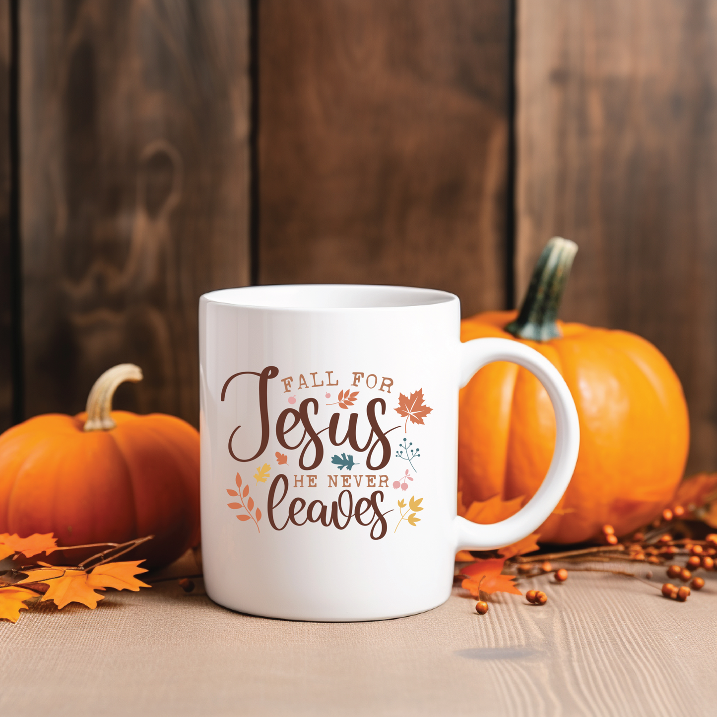 White mug with 'Fall for Jesus He Never Leaves' text, surrounded by pumpkins and leaves on a wooden surface.