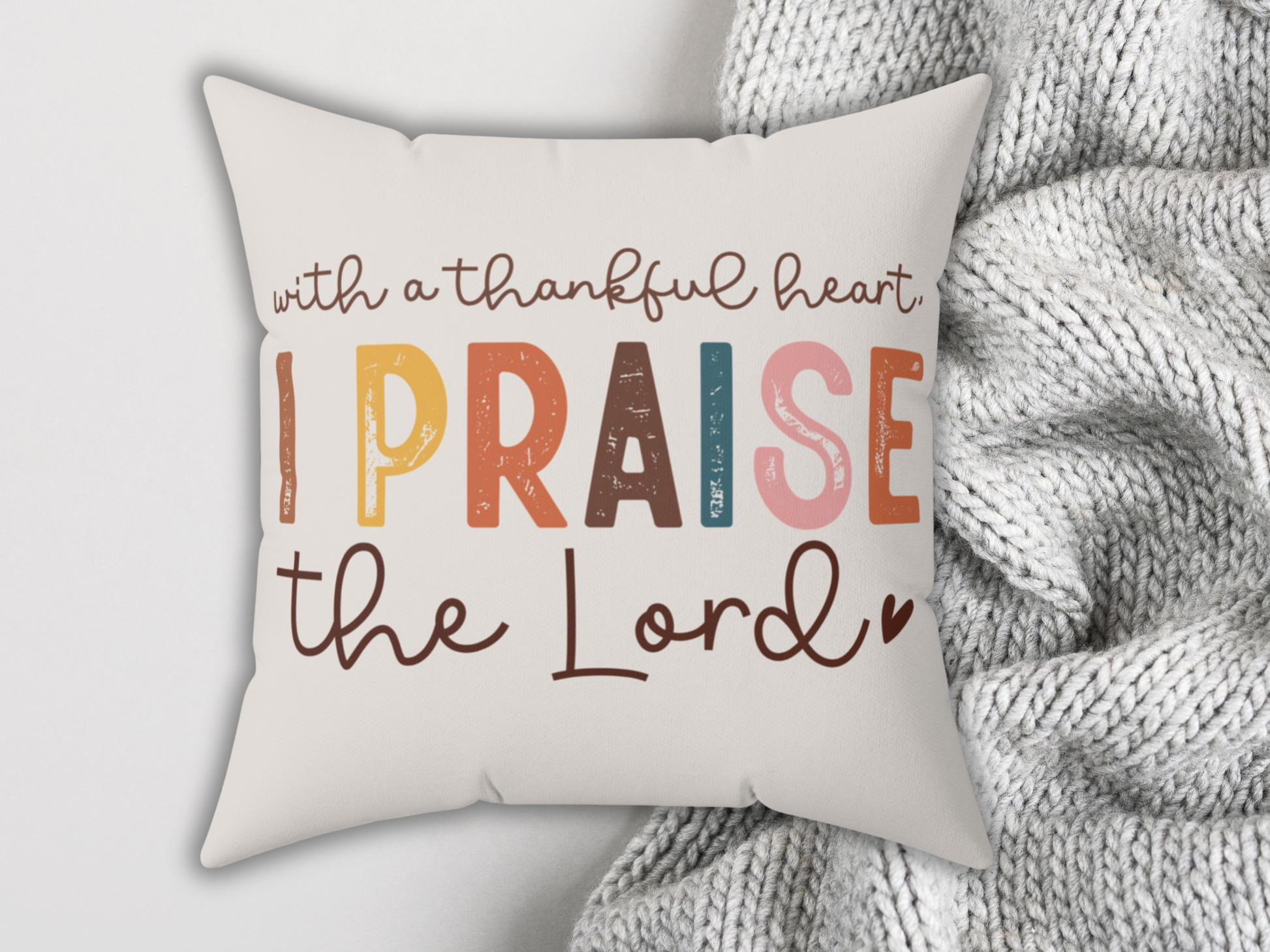 Cozy pillow with the text "with a thankful heart, I PRAISE the Lord" in colorful letters, on a gray blanket next to a latte with latte art.