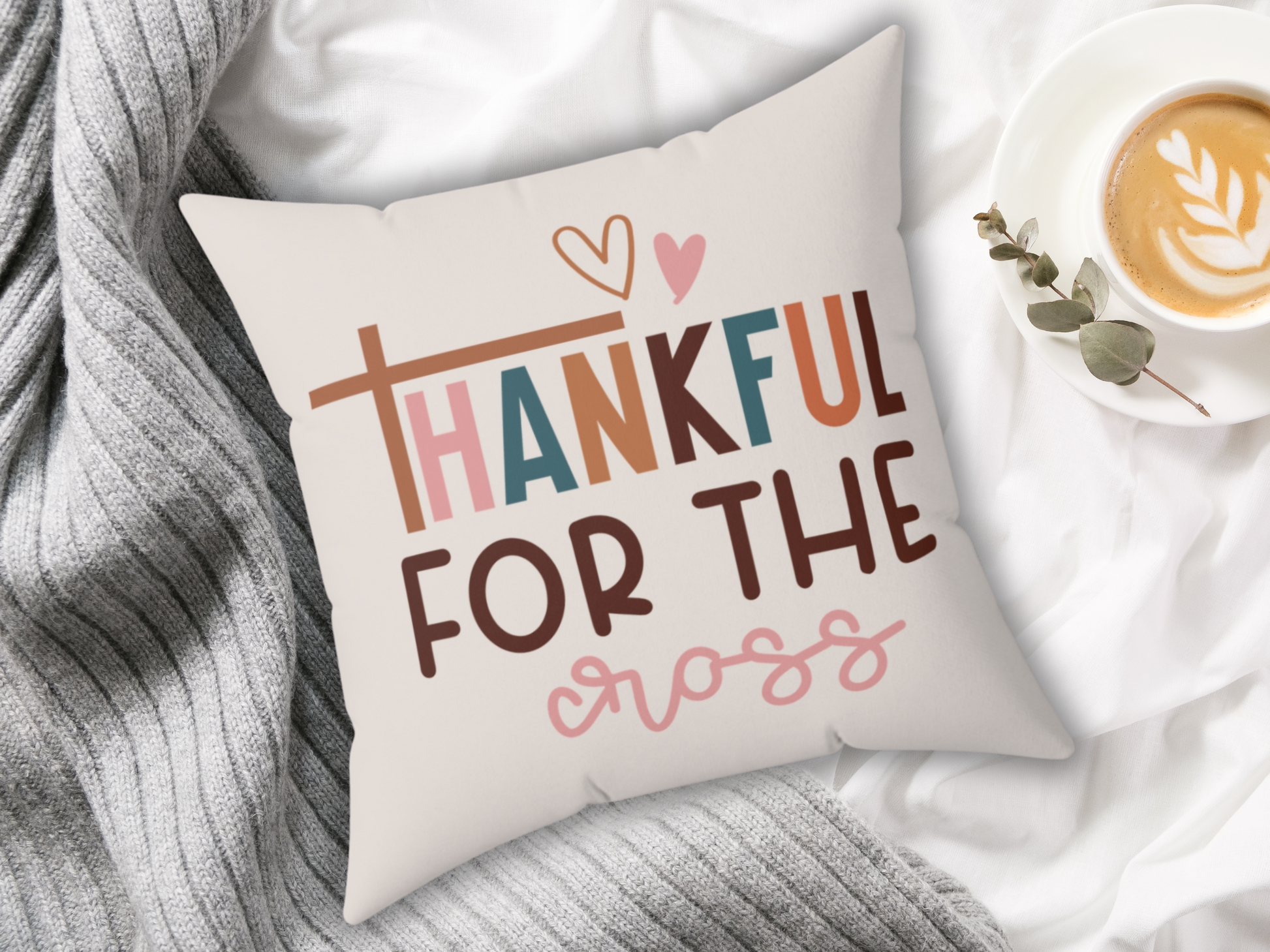 Decorative pillow on a gray blanket reads "Thankful for the cross" in colorful fonts with hearts. A latte in a cup with a leaf design sits nearby. Cozy and serene.