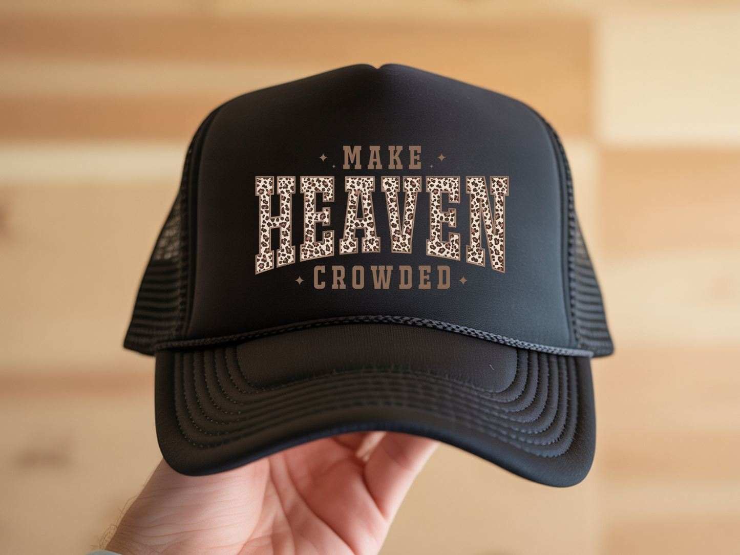 Black trucker hat with 'MAKE HEAVEN CROWDED' text held by a hand against a blurred wooden background