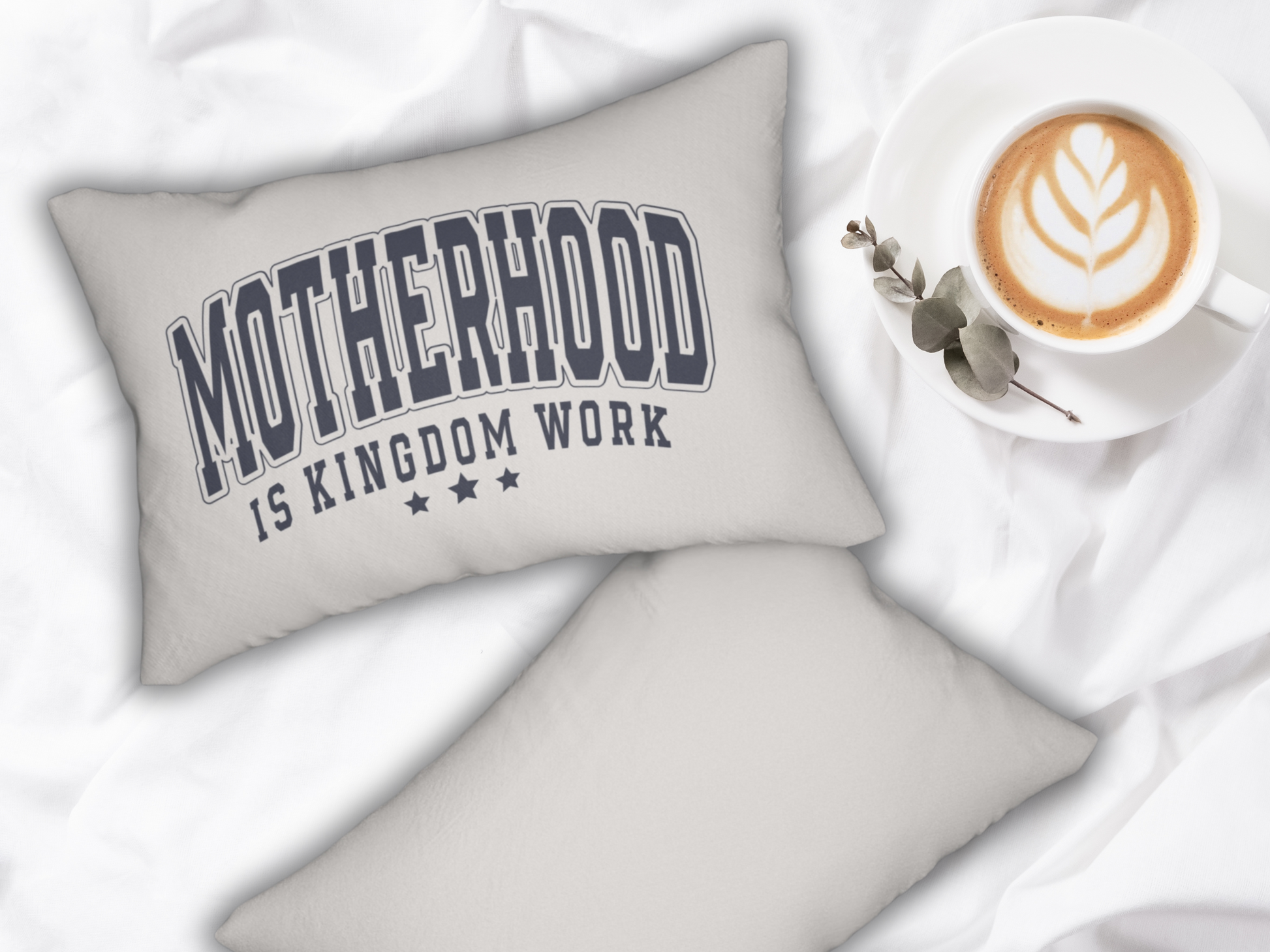 Pair of pillows on a white bedspread, one displaying the “Motherhood is Kingdom Work” design beside a latte and greenery.