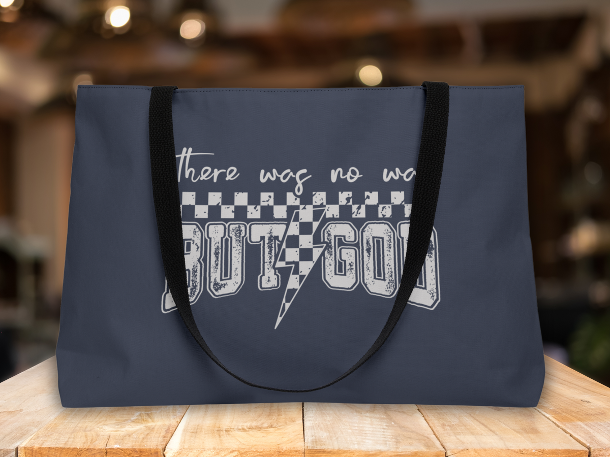 Navy blue tote bag with black straps on a wooden table, set against a blurred warm-toned indoor background, featuring “There was no way BUT GOD” text design.