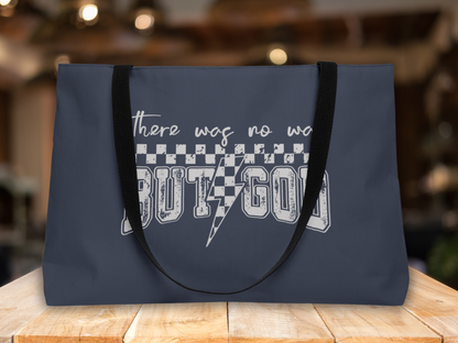 Navy blue tote bag with black straps on a wooden table, set against a blurred warm-toned indoor background, featuring “There was no way BUT GOD” text design.