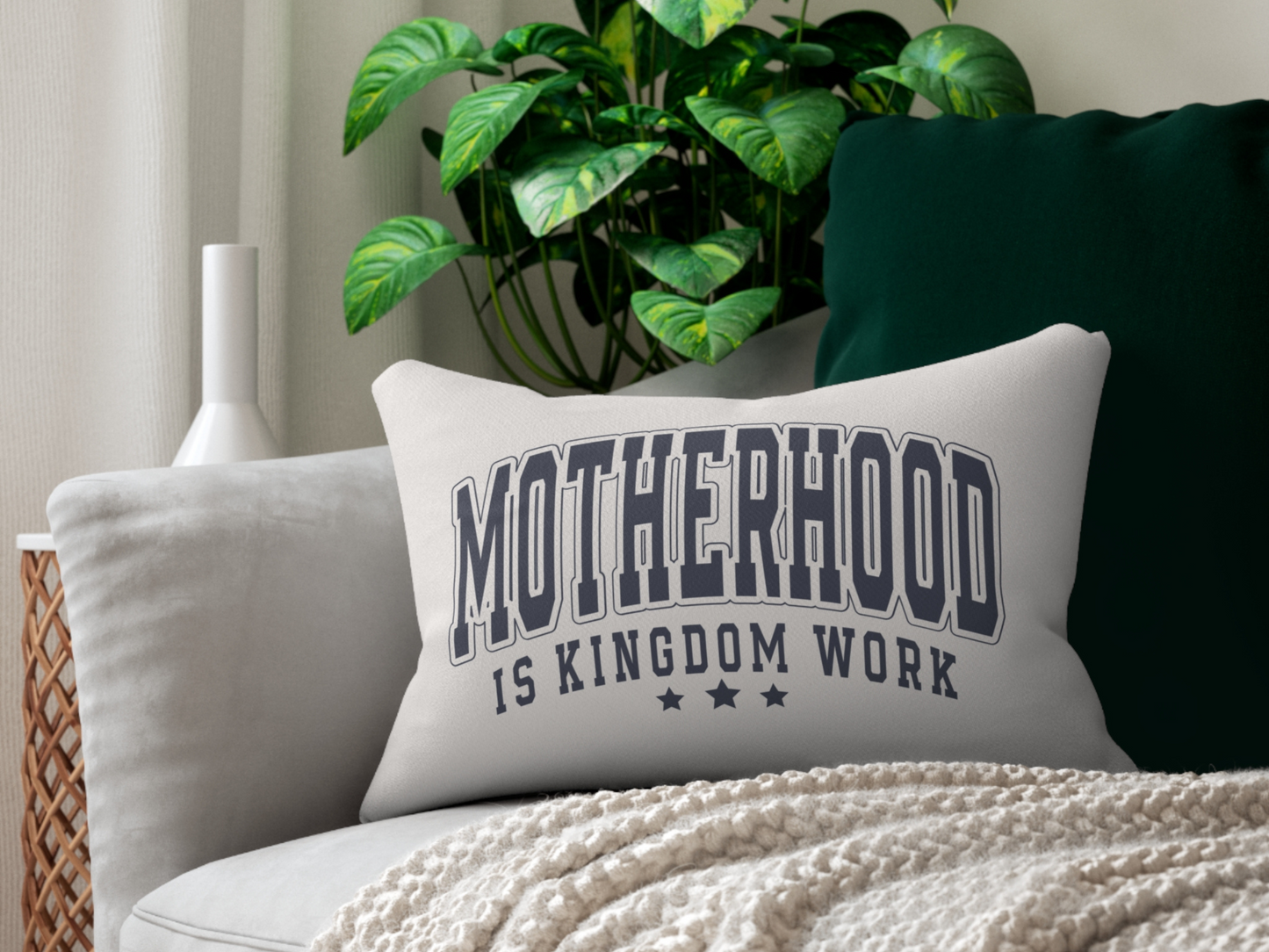 “Motherhood is Kingdom Work” pillow on a modern sofa with green cushion, throw blanket, and indoor plant.