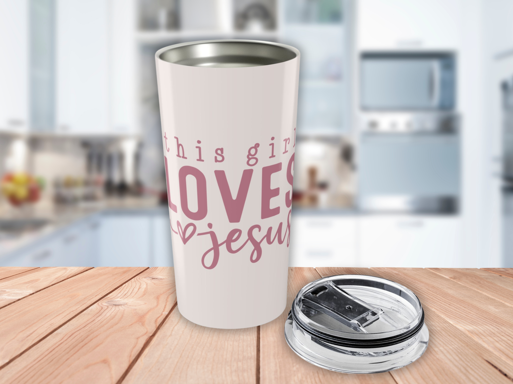 Pink tumbler with pink text 'This girl loves Jesus' on a wooden surface with a blurred kitchen background.