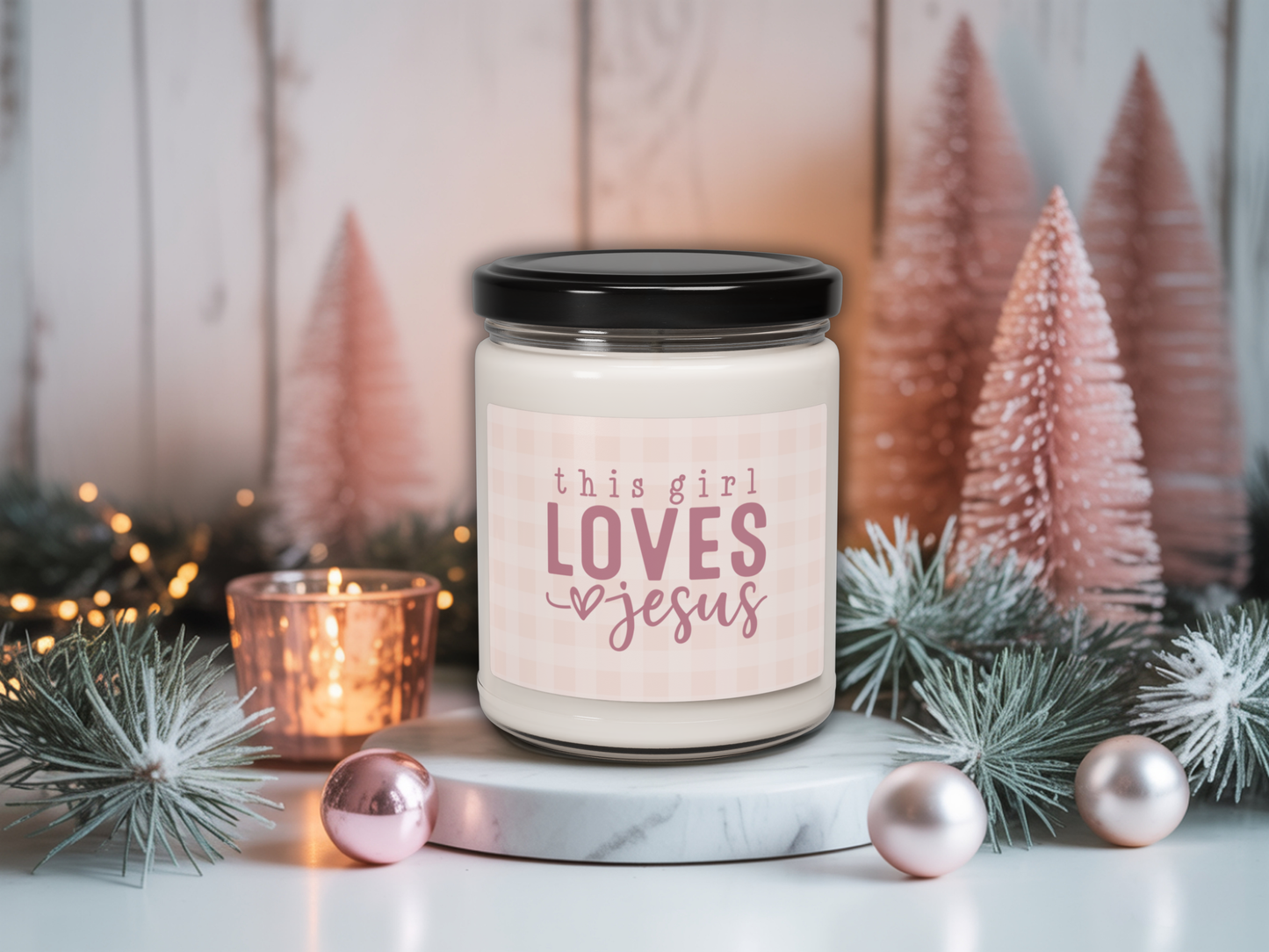 Candle jar with 'this girl loves Jesus' text in a decorative setting with Christmas trees and lights.
