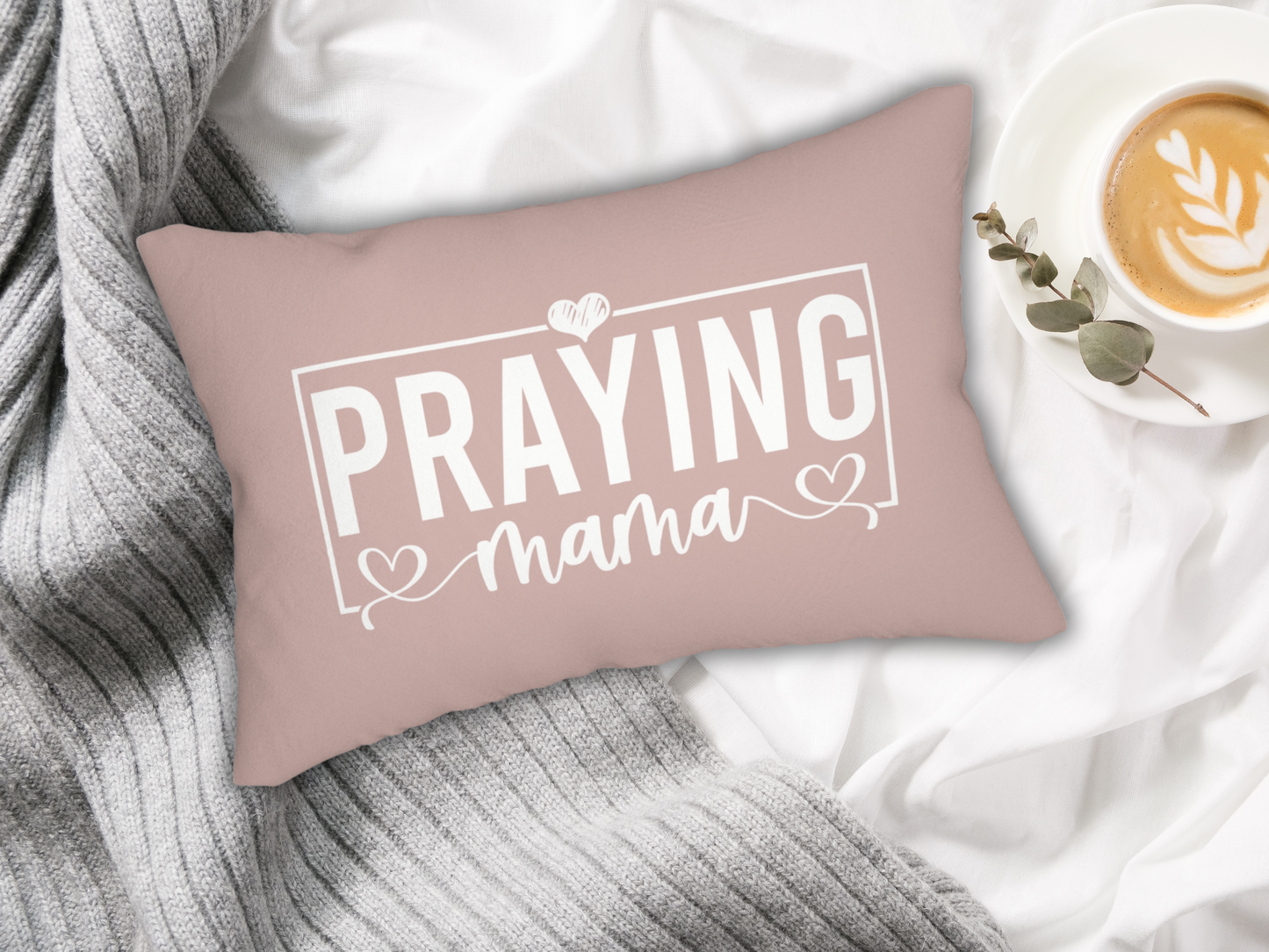 Inspirational “Praying Mama” pillow in blush pink, displayed on white bedding with gray knit blanket and latte in cup with eucalyptus sprig.